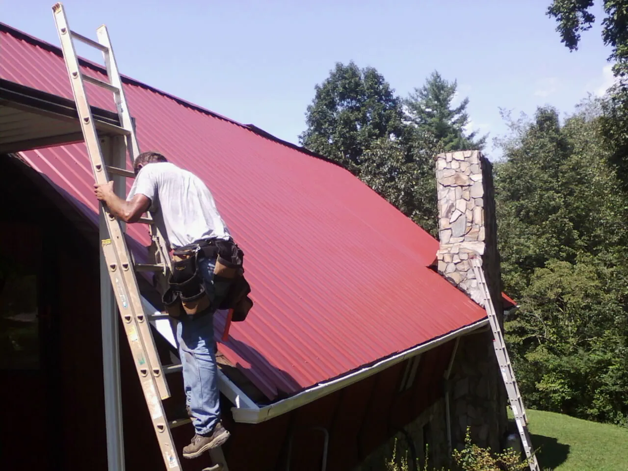 Roofer on a ladder beside a red metal roof and stone chimney — Soffit & Fascia Repair by Roofing Brothers in Grinnell